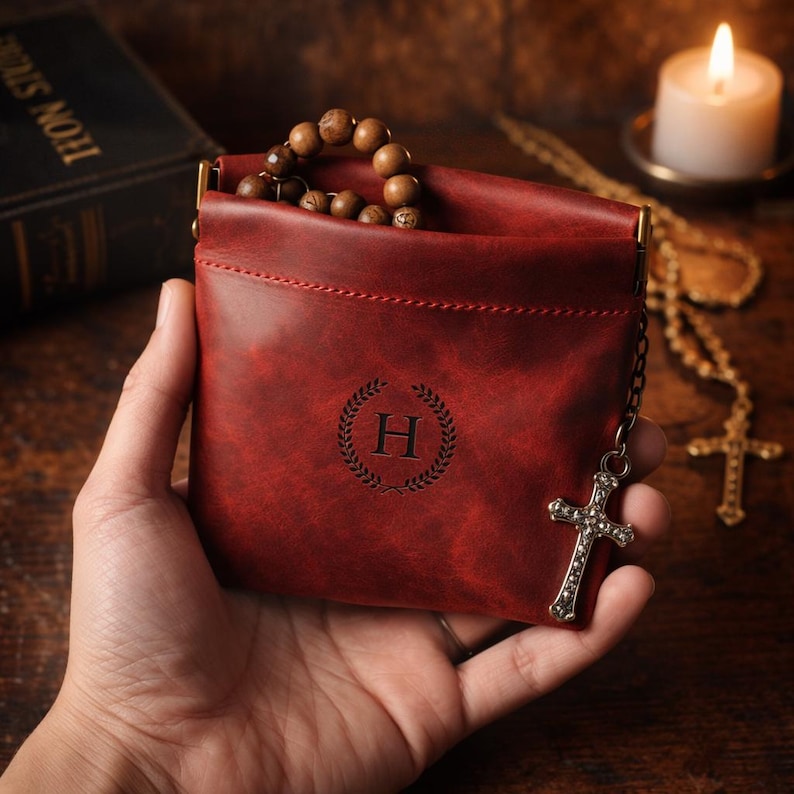 May include: A burgundy leather pouch with a decorative 'H' emblem, holding wooden beads. A silver cross charm hangs from a chain attached to the pouch. The pouch is held in a hand.