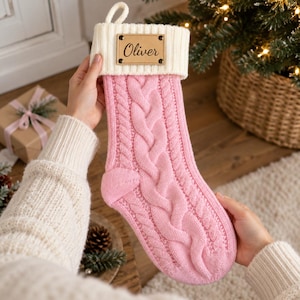 May include: A pink cable-knit Christmas stocking featuring a white ribbed cuff and a rectangular tan leather-like patch with the name Oliver engraved in black script. The stocking includes a small white loop at the top for hanging purposes.