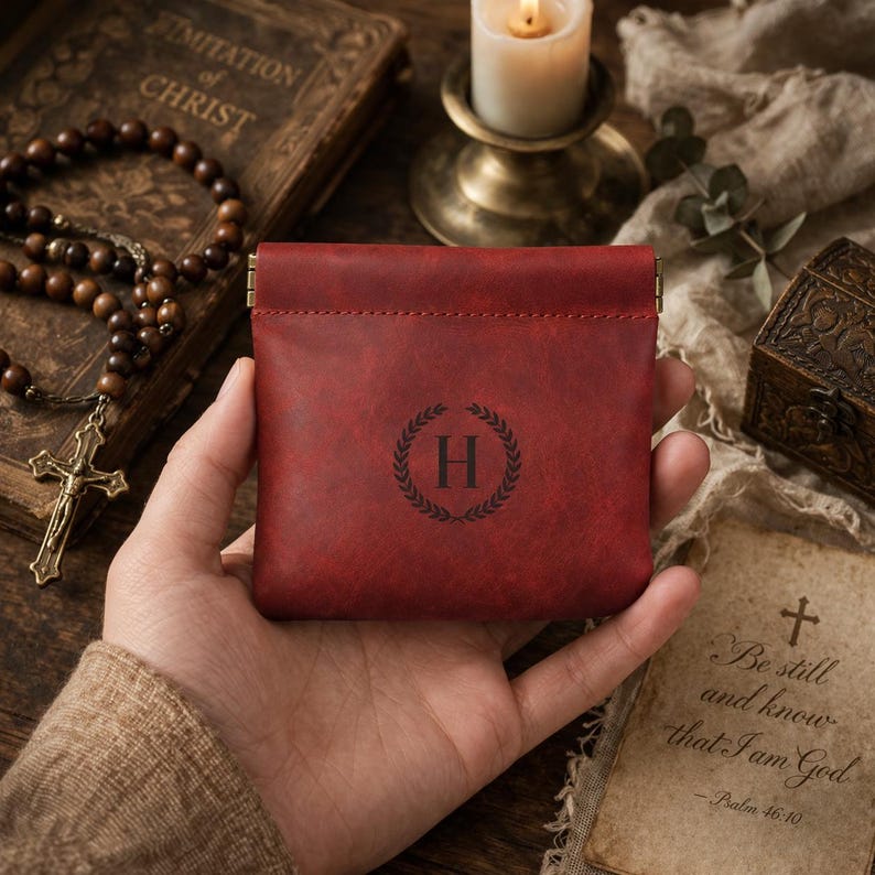May include: A red leather coin purse with a decorative wreath and the letter "H" in the center. The purse is rectangular with a clasp closure. The background includes a rosary, a candle, and a book.