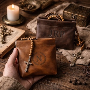 May include: Two leather pouches, one tan and one brown, are displayed with rosary beads and a cross. The tan pouch has the letter "N" inside a laurel wreath. The brown pouch has the initials "JRP". A lit candle and a book are in the background.