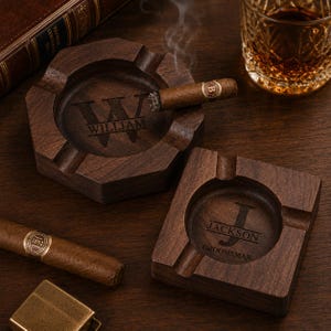 May include: Two wooden ashtrays, one hexagonal and one square, with personalized engravings. The hexagonal ashtray has "WILLIAM" engraved, and a lit cigar rests in it. The square ashtray is engraved with "JACKSON GROOMSMAN". A glass of amber liquid sits nearby.