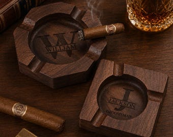 May include: Two wooden ashtrays, one hexagonal and one square, with personalized engravings. The hexagonal ashtray has "WILLIAM" engraved, and a lit cigar rests in it. The square ashtray is engraved with "JACKSON GROOMSMAN". A glass of amber liquid sits nearby.
