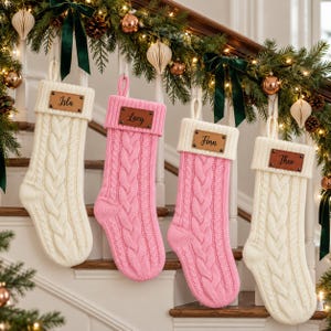 May include: Four cable-knit Christmas stockings hanging in a row. Two are cream-coloured and two are pink. Each stocking features a white cuff with a rectangular brown leather patch. The patches are engraved with the names Isla, Lucy, Finn, and Theo.
