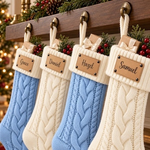 May include: Four cable-knit Christmas stockings in alternating light blue and cream colours, each featuring a white ribbed cuff, a personalised leather name tag labelled Grace, Daniel, Hazel, or Samuel, and a small gift box with greenery.