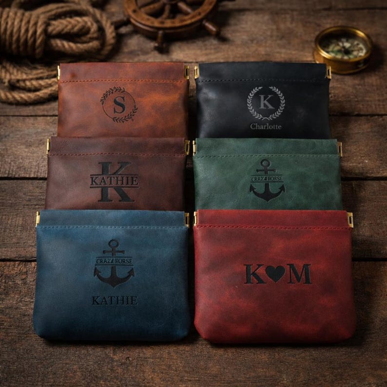 May include: A collection of leather coin purses in various colors, including brown, black, green, blue, and red. Each purse features a unique engraved design, such as initials, names, or nautical symbols. The purses have gold-colored metal accents.