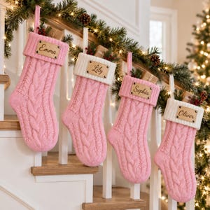 Op de afbeelding: Vier roze kabelgebreide kerstsokken met witte boorden, elk voorzien van een rechthoekig houten naamplaatje met de gegraveerde namen Emma, Lily, Sophia en Oliver. De sokken hangen op een rij en bevatten kleine cadeautjes en groen.