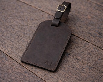 May include: A dark brown leather luggage tag with a buckle closure and a strap. The tag has the initials "AJ" embossed on the front. The tag is rectangular with rounded corners and is set on a wooden surface.