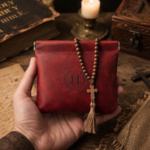 May include: A red leather pouch with a gold clasp, embossed with a letter "H" inside a laurel wreath. A wooden rosary with a cross and tassel hangs from the pouch. The pouch is held in a person's hand.