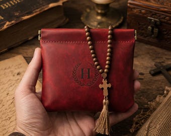 May include: A red leather pouch with a gold clasp, embossed with a letter "H" inside a laurel wreath. A wooden rosary with a cross and tassel hangs from the pouch. The pouch is held in a person's hand.
