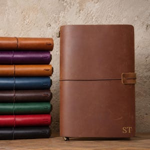 Puede incluir: Una pila de cuadernos de cuero en varios colores, incluyendo tostado, morado, azul, marrón, verde, rojo y negro, junto a un cuaderno de cuero marrón con un cierre elástico vertical y las letras ST grabadas en la parte inferior.