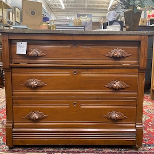 May include: A vintage wooden dresser with three drawers, featuring ornate leaf-shaped drawer pulls. The dresser has a dark brown finish and a stone top. The text "Stylish Patina" is visible on a tag on the top drawer.
