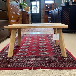 May include: A small, rectangular wooden bench with a natural finish. The bench has a slatted top and angled legs. The bench is sitting on a red patterned rug. A tag that says "Stylish Patina" is attached to the bench.