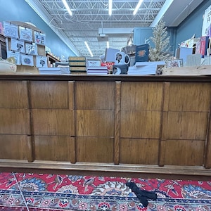 May include: A large, wooden dresser with six drawers, featuring a warm, medium-brown finish. The dresser is set on a red patterned rug and is filled with various items on top, including books and decorative objects. The overall aesthetic is vintage and rustic.