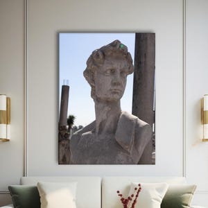 May include: A framed canvas print of a classical sculpture of a man's bust, set against a blue sky background. The sculpture is a light gray color and is displayed above a white sofa with green and white pillows. The room has modern decor.