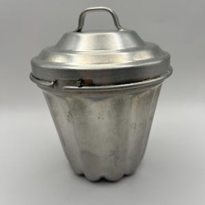 Può includere: Uno stampo vintage in metallo argentato con coperchio e manico. Lo stampo ha una forma scanalata a forma di cono e il coperchio ha una piccola maniglia ad arco. Il metallo ha un aspetto leggermente invecchiato, suggerendo che si tratta di un oggetto da cucina antico.