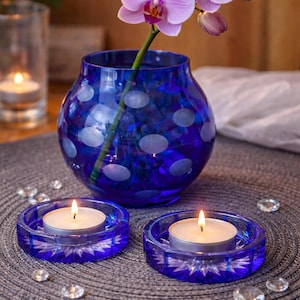 Puede incluir: Un jarrón de cristal azul con lunares blancos sostiene un tallo de orquídeas moradas. Dos candelabros de cristal azul con velas encendidas se encuentran frente al jarrón. Cristales decorativos están dispersos alrededor.