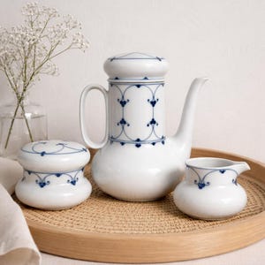 Op de afbeelding: Een witte porseleinen koffieset met blauwe bloemendetails. De set bevat een koffiepot, een melkkan en een suikerpot, allemaal gerangschikt op een ronde houten dienblad. Het ontwerp heeft een delicaat blauw patroon.
