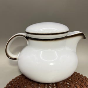 Op de afbeelding: Witte keramische theepot met een afgeronde body, een gebogen handvat en een tuit. Het deksel en de rand zijn geaccentueerd met een donkerbruine band. De theepot staat op een bruine gehaakte onderzetter. Klassiek ontwerp.
