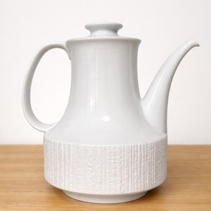 May include: A white ceramic teapot with a textured band around the base. The teapot has a curved handle, a long spout, and a lid. It sits on a wooden surface against a white background.