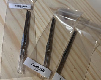 Könnte beinhalten: Drei Häkelnadeln in klarer Plastikverpackung, beschriftet mit H14, H15 und H16. Die Haken sind silberfarben und scheinen aus Metall zu sein. Der Hintergrund ist eine hellfarbige Holzoberfläche.