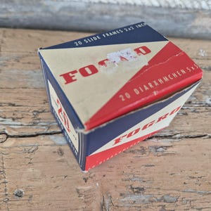 May include: A vintage box of FOGRO slide frames, 2x2 inches, with red and blue accents. The box is labeled "20 Slide Frames" and "20 Diarahmchen 5x5". The box is on a weathered wooden surface.