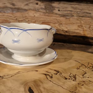 Op de afbeelding: Witte keramische soepkom met blauwe decoratieve accenten, waaronder een geschulpte rand en kleine handvatten. De kom staat op een bijpassende schotel. De achtergrond is een houten oppervlak.