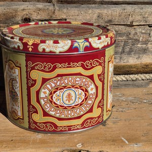 Pode incluir: Uma lata redonda vintage com um design vermelho e dourado. A lata apresenta o texto "DYAKERS PEPPARKAKOR" num oval central, rodeado por volutas e elementos decorativos. A tampa tem um padrão floral.