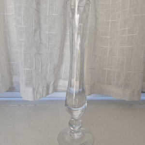 May include: A clear glass vase with a slender, elongated body and a flared top. The vase features a rounded base and a circular foot. The vase is approximately 12 inches tall and is set against a white curtain background.