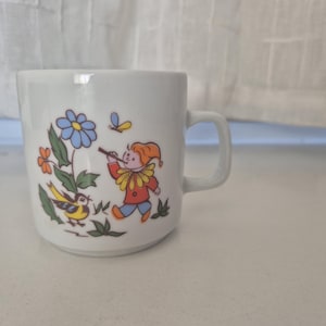Op de afbeelding: Witte keramische mok met een grillige illustratie. Het ontwerp toont een cartoon nar die fluit speelt, een blauwe bloem, een gele vogel en een vlinder. De mok heeft een eenvoudige, vierkante handgreep.