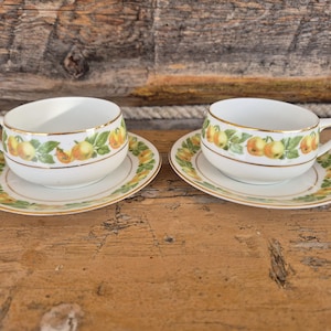 May include: Two white teacups and saucers with a fruit design. The cups and saucers have a gold trim and a pattern of green leaves and yellow and orange fruit. The teacups have handles.