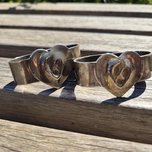 Op de afbeelding: Twee zilverkleurige servetringen met hartvormige ontwerpen. Elke ring heeft een hart in het midden, wat een vleugje romantiek toevoegt. De ringen worden gepresenteerd op een houten oppervlak, wat wijst op een rustieke of vintage esthetiek.