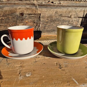 May include: Two ceramic coffee cups and saucers. One cup is white with an orange top and a matching saucer. The other cup is green with a matching saucer. The cups and saucers are on a wooden surface.