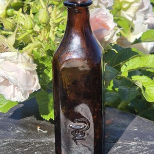 May include: A dark amber glass bottle with a narrow neck and a square base. The front of the bottle features an embossed caduceus symbol, which depicts a staff with two snakes entwined and wings at the top, centred near the bottom of the glass.