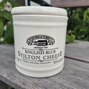May include: A white ceramic jar with a lid, featuring the text "Tuxford & Tebbutt English Blue Stilton Cheese." The jar has a black logo and text, and is sitting on a wooden surface. The jar is cylindrical and has a classic design.