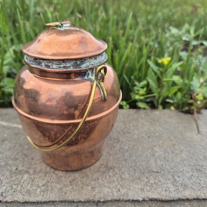 Op de afbeelding: Een kleine, ronde koperen pot met een bijpassend deksel en een dunne, gebogen messing handgreep. Het koperen oppervlak vertoont een warme, metalen glans met zichtbare tekenen van oxidatie en patina rond de rand waar het deksel het lichaam raakt.