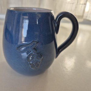 May include: A blue ceramic mug with a handle and a decorative embossed design of a hop. The mug has a glossy finish and a rounded shape, suitable for beverages.