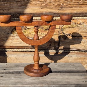 May include: A handcrafted wooden candle holder with four small bowls on top. The candle holder has a curved base and a central support. The wood has a warm, reddish-brown color, and the piece is set on a wooden surface.