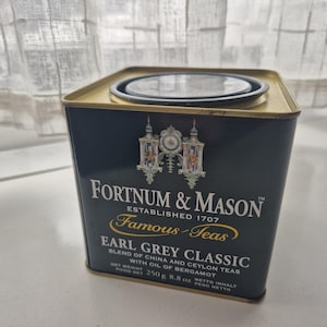 May include: A square, dark teal tea tin with a gold lid and rim. The tin features the text "Fortnum & Mason Established 1707" and "Earl Grey Classic." The net weight is 8.8 oz.