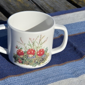 May include: A white ceramic mug with two side handles featuring a colorful illustration of three small figures with red mushroom caps sitting in green foliage. The base of the illustration includes the printed text "&copy; Elsa Beskow 1999".