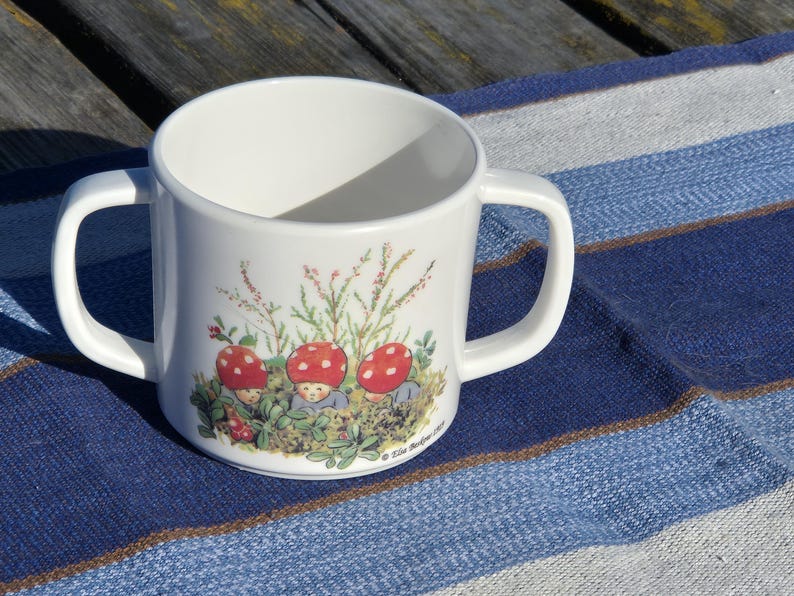 May include: A white ceramic mug with two side handles featuring a colorful illustration of three small figures with red mushroom caps sitting in green foliage. The base of the illustration includes the printed text "&copy; Elsa Beskow 1999".
