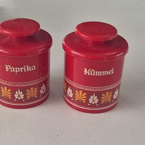 Op de afbeelding: Twee cilindrische rode kruidenpotjes met bijpassende deksels. Elk potje heeft een wit gedrukt label, waarbij er één Paprika en de andere Kümmel aangeeft. Een decoratieve band met gele en witte bloemmotieven loopt rondom de onderkant.