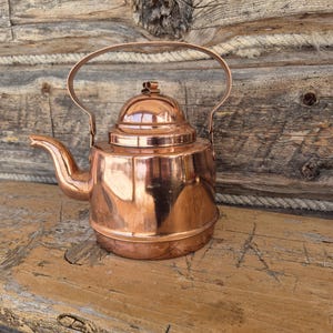 Op de afbeelding: Een glanzende koperen theepot met een gebogen tuit, een gewelfde deksel en een handvat. De ketel staat op een houten oppervlak tegen een rustieke houten achtergrond. Het koper reflecteert de omgeving.