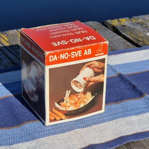 以下が含まれることがあります： DA-NO-SVE ABマルチグレーターの正方形の段ボール製製品ボックス。箱は主に赤と白で、DA-NO-SVEおよびMulti-Graterという文字が印刷されています。前面パネルには、白い円筒形のキッチン用グレーターの写真イラストが描かれています。
