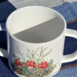 May include: A white ceramic mug with two handles featuring a colorful illustration of three small figures with red mushroom caps on their heads, surrounded by green foliage and small pink flowers, with the text Elsa Beskow 1919 printed at the base.