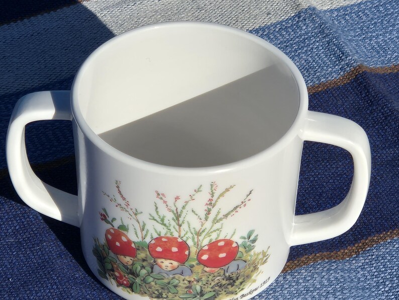 May include: A white ceramic mug with two handles featuring a colorful illustration of three small figures with red mushroom caps on their heads, surrounded by green foliage and small pink flowers, with the text Elsa Beskow 1919 printed at the base.