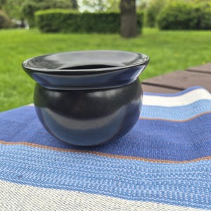 Op de afbeelding: Een zwarte keramische kom met een brede rand en een afgeronde basis. De kom staat op een blauw-wit gestreepte stof, met een groene, grasrijke achtergrond. De kom heeft een glanzende afwerking en een eenvoudig, elegant ontwerp.