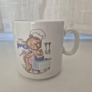Könnte beinhalten: Weiße Keramiktasse mit einem Cartoon-Bärenkoch, der eine Kochmütze und eine Schürze trägt und an einem Herd kocht. Der Bär lächelt. Die Tasse hat einen gebogenen Griff und ein schlichtes Design.