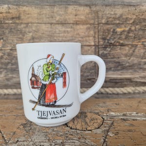 Może przedstawiać: Biały ceramiczny kubek z ilustracją narciarza w tradycyjnym stroju, trzymającego kijki narciarskie, z napisem "TJEJVASAN OXBERG - MORA 30 Km". Kubek ma zakrzywiony uchwyt i okrągły wzór w kolorach czerwonym, zielonym i białym.
