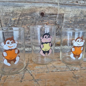 Puede incluir: Tres vasos de vidrio transparente con ilustraciones de monos de dibujos animados. Cada mono es marrón, negro y blanco, sosteniendo un objeto naranja en forma de corazón. Los vasos son cilíndricos y están sobre una superficie de madera.