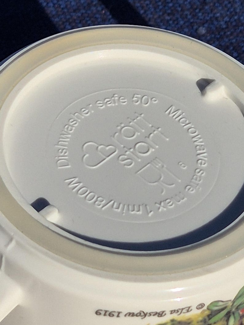 May include: The underside of a white ceramic mug featuring embossed text that reads R&auml;tt Start, Dishwasher safe 50 degrees, Microwave safe max 1 min/800W, and a small icon of a fork and glass. The base also shows the text Elsa Beskow 1919.